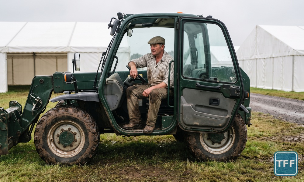 A farmer in a flat cap sits alone in the open cab of a plain dark green telehandler on a quiet agricultural showground trade stand, white marquees behind
